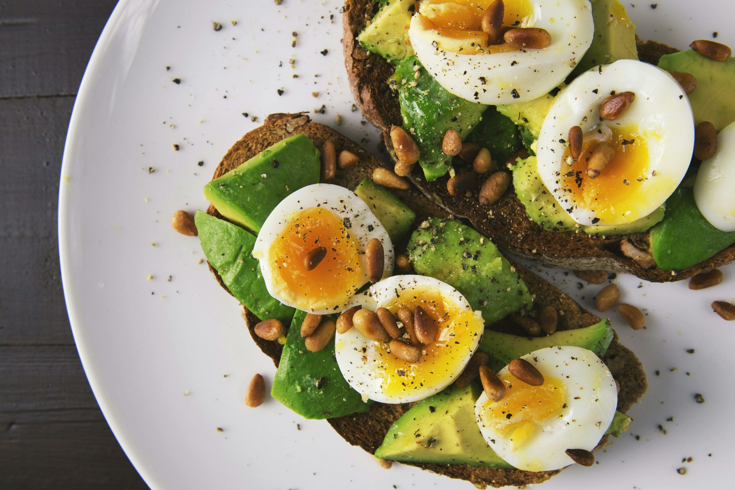 Home Delicious avocado toast topped with soft-boiled eggs and pine nuts on a white plate.