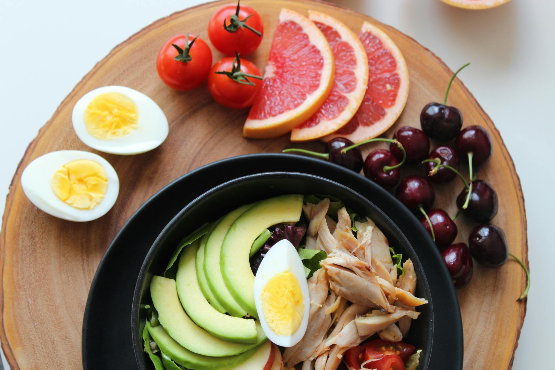Home A vibrant, healthy salad bowl with avocado, eggs, tomatoes, grapefruit, and cherries on a wooden board.